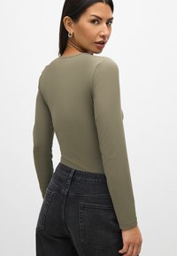 Vihreä oliivinvärinen pitkähihainen bodysuit, jossa on tiukka muotoilu ja pyöreä pääntie, yhdistettynä tummanharmaisiin korkeavyötäröisiin farkkuihin.