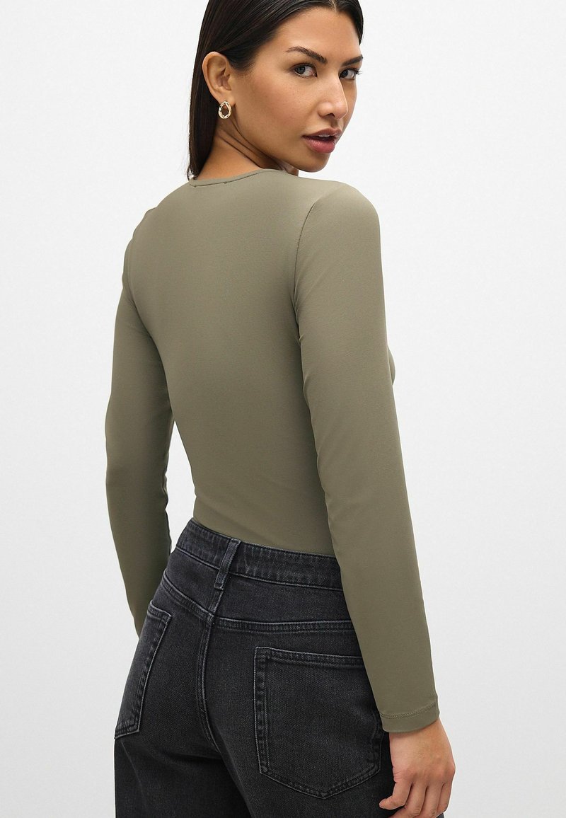 Vihreä oliivinvärinen pitkähihainen bodysuit, jossa on tiukka muotoilu ja pyöreä pääntie, yhdistettynä tummanharmaisiin korkeavyötäröisiin farkkuihin.