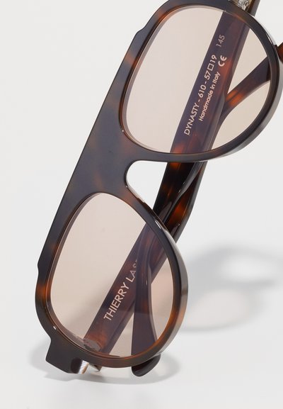Occhiali da sole tartarugati con grandi montature angolari marroni e lenti di un marrone chiaro. Presentano il marchio "THIERRY LASRY" sulle astine.