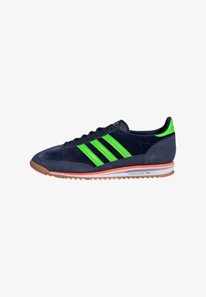 Shoe atlético em camurça azul-marinho com três riscas verdes ousadas, sola laranja e entressola branca. Apresenta um perfil baixo e uma superfície texturizada.