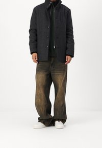 Šedý kabát z vlněné směsi s vysokým límcem, zapínáním na knoflíky a kapsou na hrudi, v kombinaci s béžovými širokými džínami a bílými teniskami.