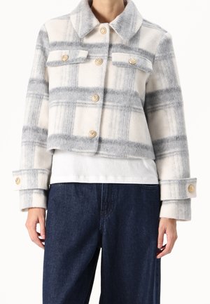 Kropskåret hvid og grå ternet jakke med guldknapper, brystlommer med klapper og brede manchetter, kombineret med mørkeblå jeans og en hvid skjorte.