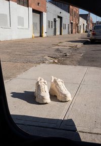 Paire de baskets beiges placée sur un trottoir en ville près de bâtiments industriels sous une lumière du jour claire.