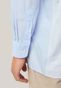 Chemise boutonnée rayée bleu clair à manches longues, dotée de boutons couleur perle aux poignets. Le tissu semble lisse et soigné.