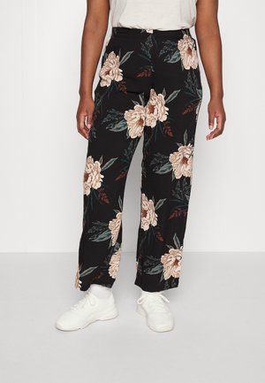 Personne portant un pantalon large noir à motif floral beige, des baskets blanches et une chemise claire rentrée, debout sur un sol clair.