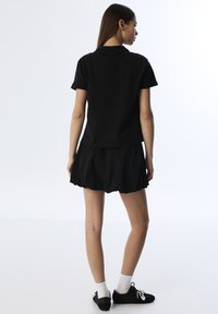 Camiseta polo de algodón negra con mangas cortas, combinada con una falda negra acampanada. Modelo con zapatos negros y calcetas blancas. Diseño minimalista.