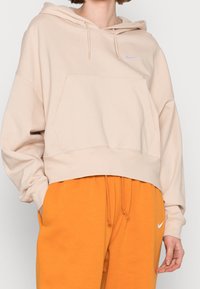 Beige oversized hoodie with drawstrings, front kangaroo pocket, and small white Nike logo. Paired with bright orange sweatpants.