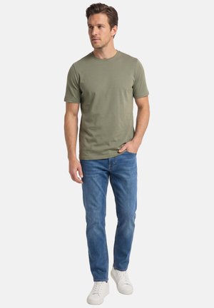 Man draagt een olijfgroen T-shirt met korte mouwen, blauwe jeans en witte sneakers, staat met één hand in de zak tegen een witte achtergrond.