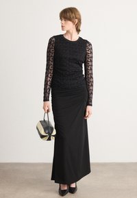 Schwarzes Kleid mit langen, durchsichtigen, gemusterten Ärmeln. Raffung im taillierten Schnitt und einem bodenlangen Rock. Die Modells hält eine Handtasche in Schwarz und Creme.