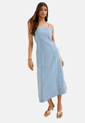 Donna che indossa un vestito midi in denim azzurro chiaro senza maniche con bretelle sottili e sandali bianchi con tacco, in piedi davanti a uno sfondo semplice.