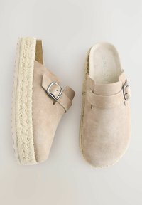 Zuecos de gamuza beige con tacón de cuña, que presentan una suela de yute trenzado y un detalle de hebilla plateada en el lateral, ofreciendo una textura suave.