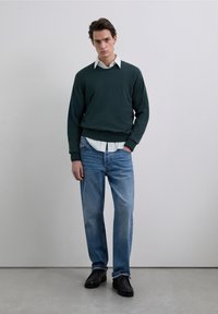 Mörkgrön tröja över en ljusrandig skjorta, kombinerad med ljusblå jeans och svarta skor. Modellen står mot en neutral bakgrund.