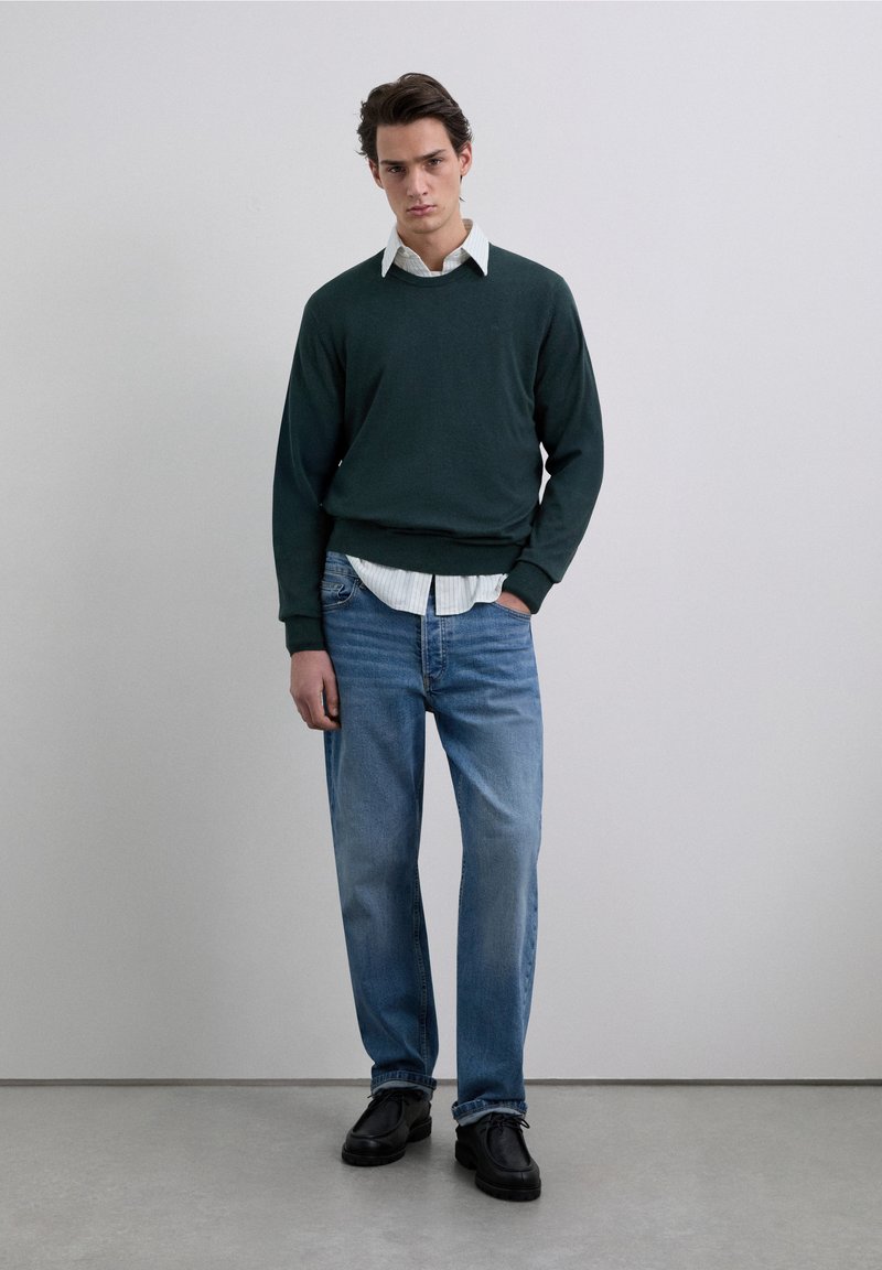 Mörkgrön tröja över en ljusrandig skjorta, kombinerad med ljusblå jeans och svarta skor. Modellen står mot en neutral bakgrund.