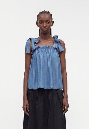 Femme aux cheveux tressés portant un haut bleu sans manches à bretelles nouées et une jupe noire taille haute, debout devant un fond blanc uni.