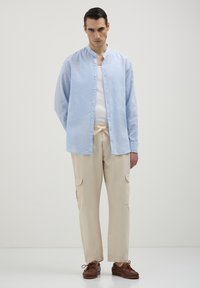 Camicia azzurra chiara con colletto alla cinese, abbinata a pantaloni cargo beige con tasche laterali e scarpe stringate marroni.