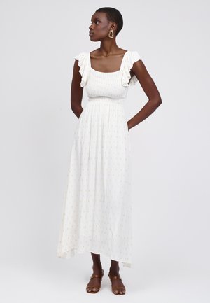 Weißes Maxikleid mit quadratischem Ausschnitt, gerafften Trägern und einem gerafften Oberteil. Mit dezenten goldenen Mustern durchgehend. Braune Sandalen.