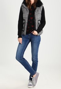 Polstret vest i sort og grå med en pelsforet hætte. Båret over en sort sweater med røde hjerter, sammen med blå jeans og grå sneakers.