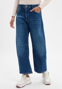 Persona che indossa jeans blu larghi e sneakers bianche chunky, con una mano in tasca su uno sfondo semplice.