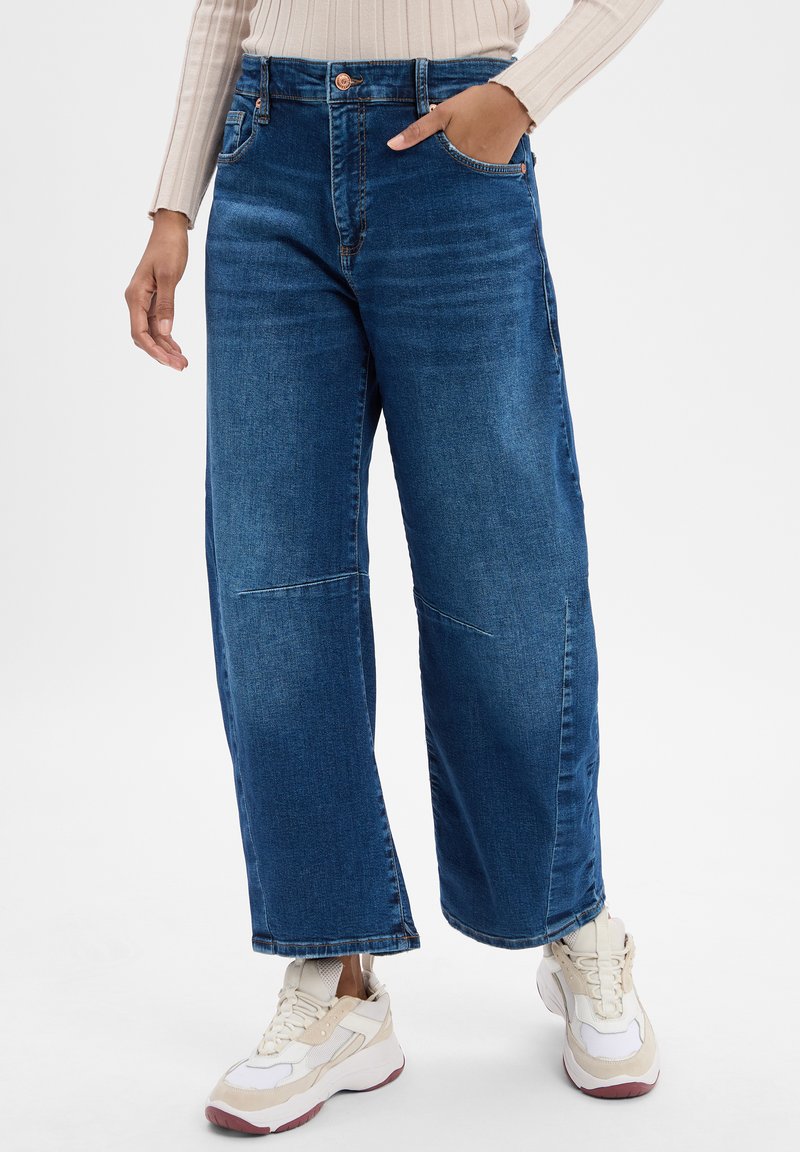Persona che indossa jeans blu larghi e sneakers bianche chunky, con una mano in tasca su uno sfondo semplice.