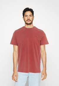 T-shirt di cotone rosso con maniche corte, scollatura a girocollo e un sottile dettaglio del logo. Texture leggermente sbiadita e vestibilità rilassata. Indossato con jeans azzurri chiari.