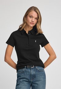 Camisa polo negra de manga corta, con tapeta de botones y pequeño logotipo bordado. Combinada con unos jeans rectos clásicos de color azul.