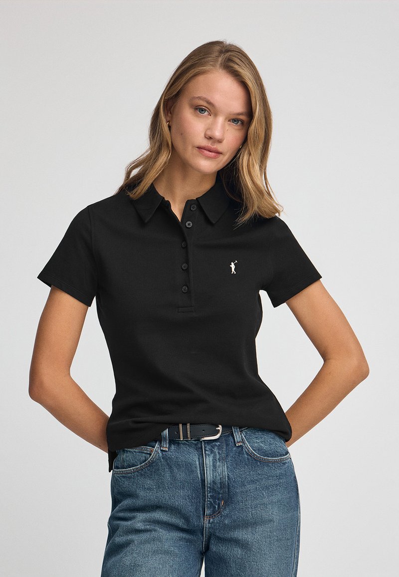 Camisa polo negra de manga corta, con tapeta de botones y pequeño logotipo bordado. Combinada con unos jeans rectos clásicos de color azul.
