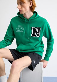 Green Nike hoodie with embroidered "N" logo, white detailing, adjustable drawstring, and a front pocket. Worn with black athletic shorts.