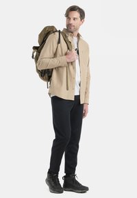Camicia beige a maniche lunghe con bottoni, T-shirt bianca, pantaloni neri a sigaretta e scarponi da trekking neri; zaino verde oliva a tracolla.