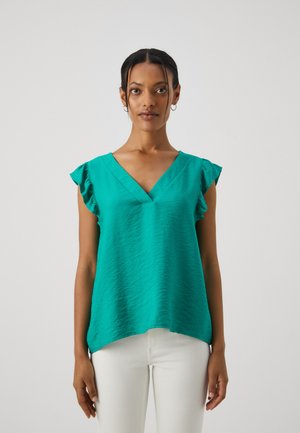 Top senza maniche di colore teal con scollatura a V, caratterizzato da spalline a volant e un tessuto leggero e texturizzato. Abbinato a pantaloni bianchi.