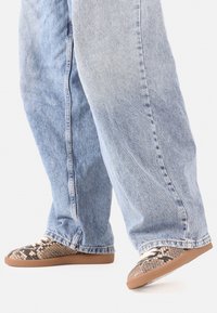 Pantofi slip-on cu un model de imprimeu de șarpe, culori maro și negru, partea superioară texturată și talpă flexibilă din cauciuc, asortați cu blugi de culoare albastru deschis.