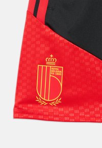 Rotes Sporttrikot mit tonalem Rautenmuster, versehen mit einem gelben Wappen mit der Aufschrift "Royal Belgian FA 1895" und schwarzen Akzenten auf den Schultern.