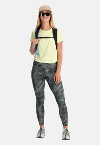 Maglietta gialla chiara a maniche corte, leggings grigi con motivo astratto verde, zaino nero, occhiali da sole e cappellino beige. Tessuto strutturato, vestibilità atletica.