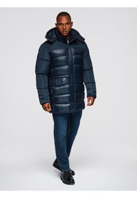 Hombre caminando hacia adelante con un abrigo largo acolchado de invierno azul marino, jeans azules y botas negras con cordones, en un fondo gris liso.