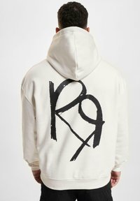 Creme farvet hættetrøje med et stort sort grafisk logo på ryggen. Har snore i hætten og ribbede manchetter. Fremstillet af blødt bomuldsstof.