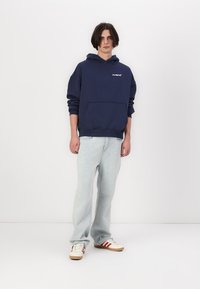 Felpa navy con tasca frontale e logo, abbinata a jeans azzurri e sneakers con motivo che presentano accenti rossi e beige.