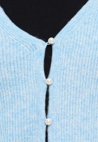 Ανοιχτό μπλε πλεκτή ζακέτα με V-λαιμόκοψη, ριμπ υφή και τρία μαργαριταρένια κουμπιά στο μπροστινό μέρος. Μαύρο ύφασμα ορατό από κάτω.