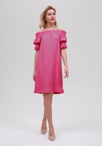 Off-shoulder rosa klänning med puffärmar, texturerad tyg och rak skärning, matchad med nude-klackar mot en neutral bakgrund.