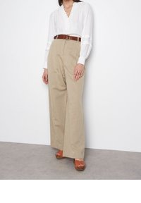 Kvinna iförd beige vidbyxor, vit långärmad blus, brun bälte, bruna öppna klacksandaler och guldringar på vänster hand.