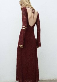Burgundy lace dress with long sleeves and an open back. Features gold accents on the sleeves and a tie closure at the neck.