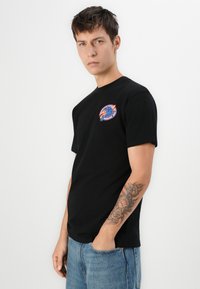 Zwarte katoenen T-shirt met korte mouwen en ronde hals, voorzien van een kleurrijke grafische print op de linkerborst. Gemaakt met blauwe denim jeans.
