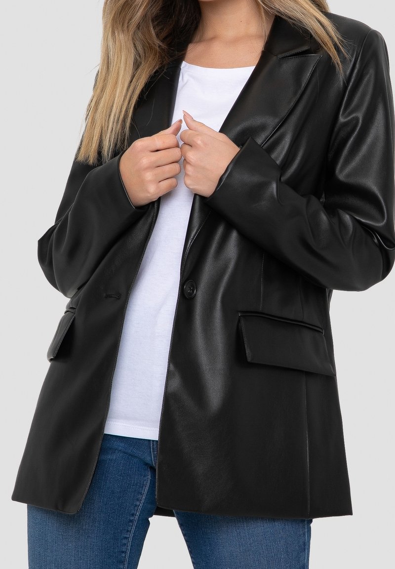 Personne portant un blazer en cuir noir sur une chemise blanche et un jean bleu, ajustant le revers du blazer avec les deux mains.