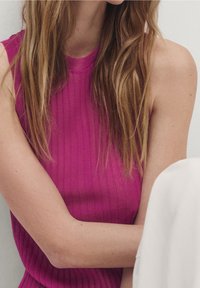 Jasnoróżowy prążkowany top bez rękawów z wysokim kołnierzem, o wyczuwalnej fakturze i luźnym kroju, zestawiony z zakrytymi ramionami i neutralnym tłem.