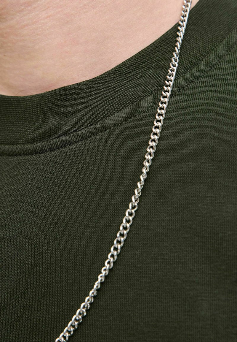 T-shirt en coton vert avec un col côtelé, arborant un collier en chaîne argentée brillante sur un fond en tissu texturé.