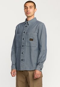 Camisa de botones de denim azul claro. Presenta dos bolsillos frontales, un cuello y una etiqueta en el lado izquierdo del pecho. Mangas largas con puños de botón.