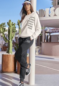 Felpa crema con testo nero; pantaloni jogger neri con ricamo bianco; sneakers casual; ambiente esterno con cactus e architettura moderna.