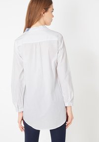 Camisa blanca de manga larga con dobladillo redondeado, pliegue en la parte trasera y un cuello con acento azul. Tela suave con un diseño ajustado.