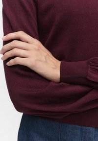 Burgundowa sweter z dzianiny z prążkowanymi mankietami. Zbliżenie na dłoń spoczywającą na rękawie, ukazujące miękką fakturę i szczegółowe szycie.