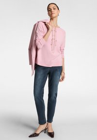 Maglione rosa con maniche lunghe, scollatura rotonda e collana dorata a strati. Jeans scuri e décolleté marroni. Su uno sfondo neutro.