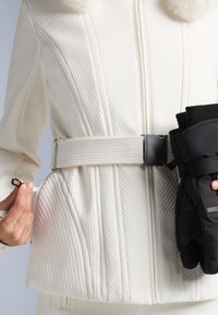 Jachetă albă matlasată cu guler din blană sintetică, având o curea orizontală lată și textură verticală cu nervuri; ținând mănuși negre cu un freamăt.