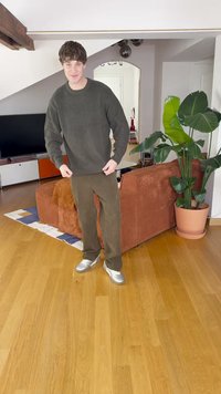 Pulover verde cu dungi, pantaloni de catifea maro și adidași argintii purtați în interior. Prezintă un pardoseală din lemn și o canapea texturată portocalie cu plante.
